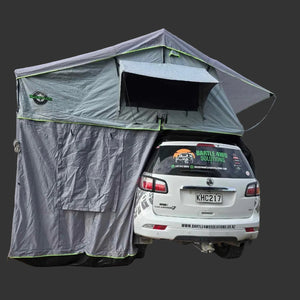 A gray rooftop tent with green trim is mounted over the rear of a white SUV on a brick driveway in front of a modern building.